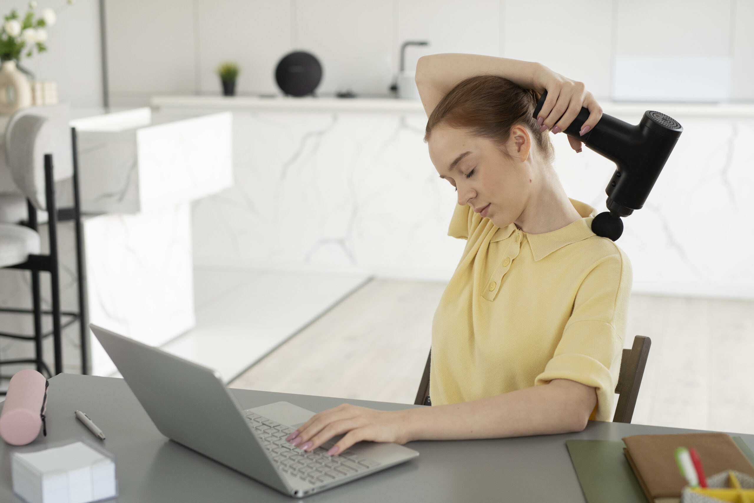 Woman at her laptop using a massage gun to work tension out of her neck, a metaphor for addressing tension in working relationships.