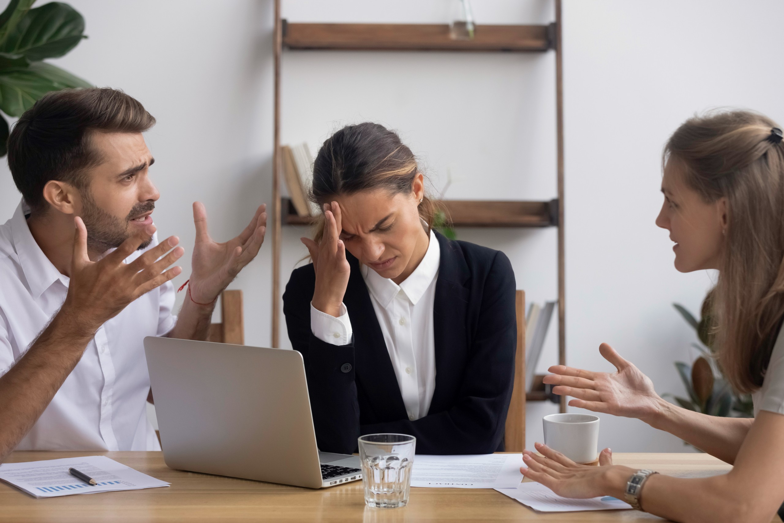 Frustrated woman rubbing her forehead between two coworkers arguing, like a customer caught between teams that don't communicate