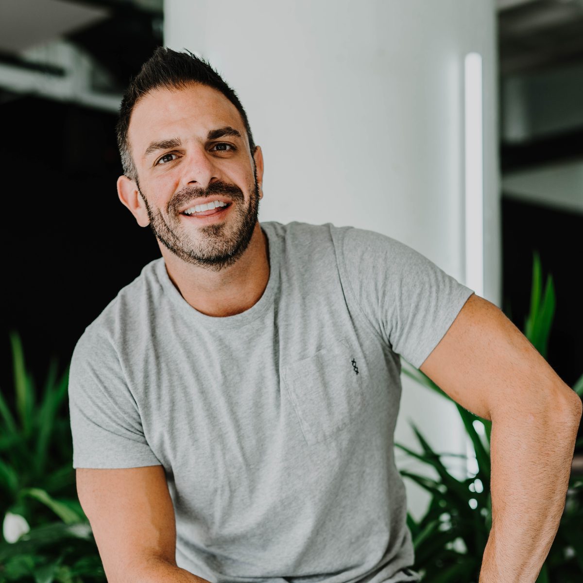 Josh Sample, founder of Drive Social Media, smiling in a grey T-shirt in a bright studio with plants behind him